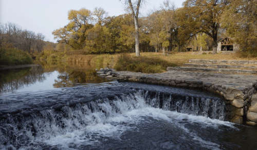 A serene river scene with a small waterfall, surrounded by autumn trees and a rustic shelter in the background.