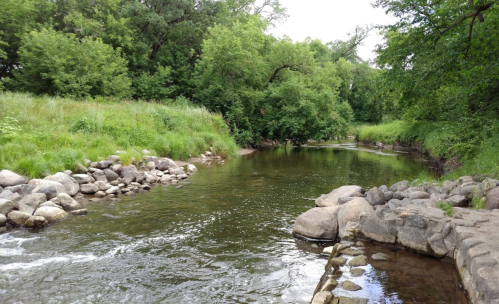 A serene river flows through a lush green landscape, lined with rocks and trees under a cloudy sky.