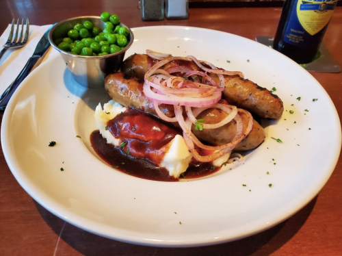 A plate with sausages topped with onions, served with mashed potatoes, gravy, and a side of green peas.