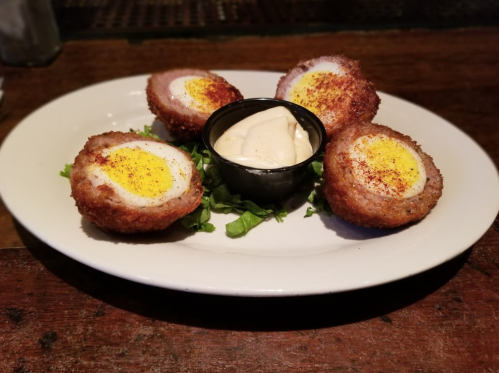 A plate of five crispy Scotch eggs served with a small bowl of dipping sauce on a bed of greens.