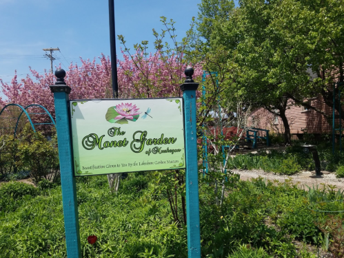 Sign for the Monet Garden of Muskegon, surrounded by blooming trees and lush greenery on a sunny day.