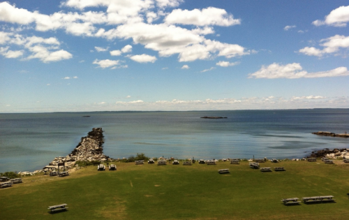 A serene view of a calm lake with a rocky shoreline and picnic tables on a grassy area under a blue sky with clouds.