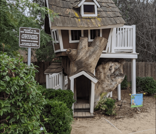 A whimsical treehouse with a wooden structure, nestled among greenery, featuring a sign that welcomes visitors.
