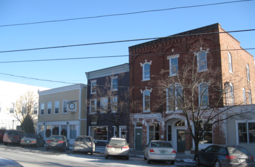 A street view of two historic buildings with shops, parked cars, and clear blue sky.