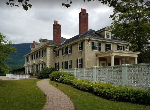 A large, elegant house with multiple chimneys, surrounded by greenery and a white fence, set against a mountainous backdrop.