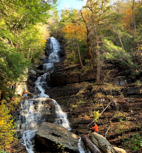 A tall waterfall cascades down rocky cliffs, surrounded by vibrant autumn foliage and two people near the base.