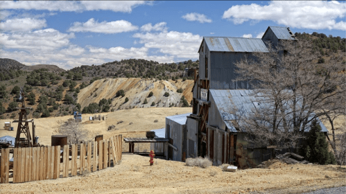 A weathered mining building stands in a barren landscape, surrounded by hills and scattered trees under a cloudy sky.