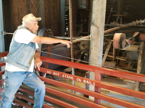 A man in a denim vest and cap gestures while standing by a railing in a rustic, industrial setting.