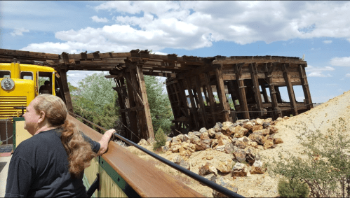 A person stands near a dilapidated wooden structure on a hillside, with rocks and a yellow train in the background.