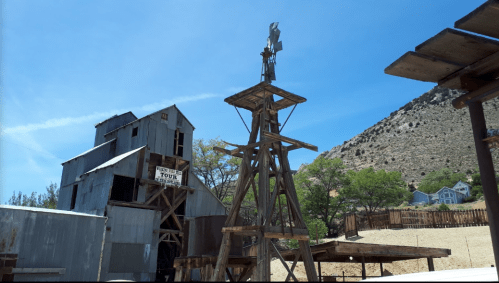 A wooden windmill stands in front of a rustic building against a clear blue sky and mountainous backdrop.