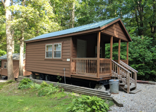 A small brown cabin with a green roof, surrounded by trees, featuring a porch and steps leading to the entrance.