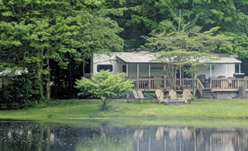 A serene lakeside scene featuring a trailer home surrounded by lush greenery and a calm water reflection.