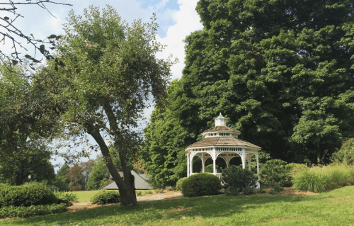 A white gazebo surrounded by lush greenery and trees under a partly cloudy sky.