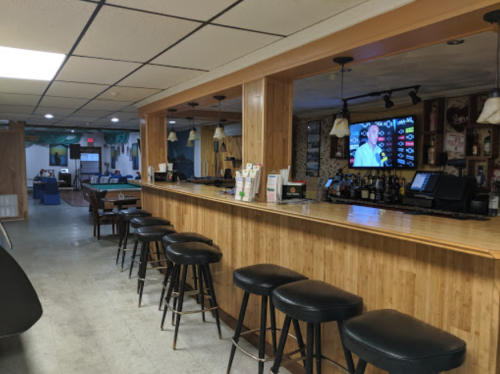 A wooden bar with black stools, a TV displaying a menu, and a spacious lounge area in the background.