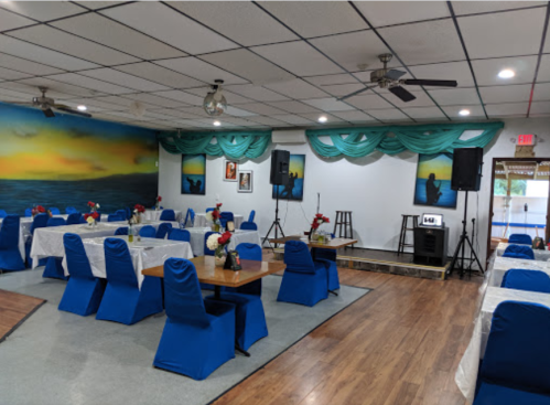 A spacious dining area with blue chairs, white tablecloths, and floral centerpieces, featuring a stage and colorful wall art.