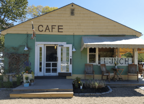 A quaint café with a green exterior, wooden deck, and outdoor seating under a canopy, featuring a sign that says "LUNCH."
