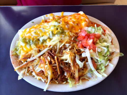 A plate of enchiladas topped with green and yellow cheese, served with shredded lettuce, tomatoes, and crispy potatoes.