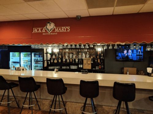 A bar area with a countertop, stools, shelves of liquor, and a TV, featuring the sign "Jack & Mary's Restaurant."