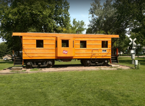 An orange train car on display in a grassy park, surrounded by trees and a railroad crossing signal nearby.