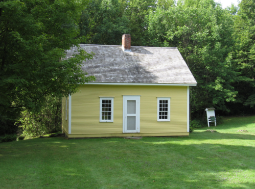 A small yellow house with a sloped roof, surrounded by green trees and grass. A white chair is visible nearby.