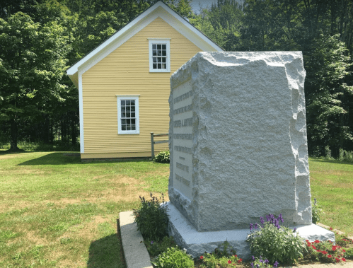 A granite monument surrounded by flowers, with a yellow house and trees in the background on a sunny day.