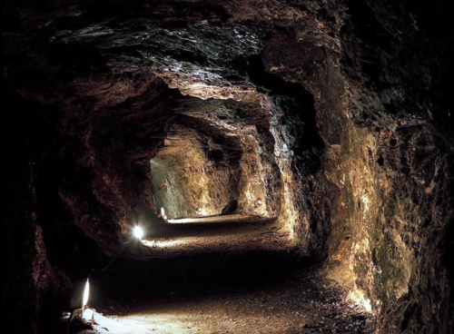 A dimly lit underground tunnel with rocky walls and a dirt path, illuminated by small lights along the sides.