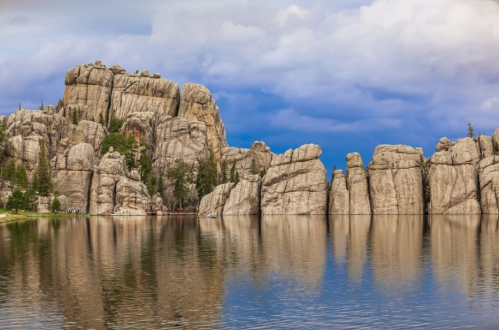 Rocky cliffs reflect in a calm lake under a blue sky with clouds, surrounded by trees.