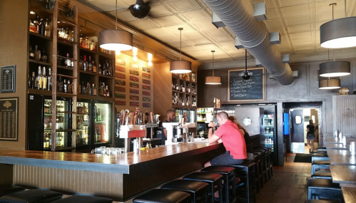 A cozy bar interior with a wooden counter, shelves of liquor, and patrons seated at the bar enjoying drinks.