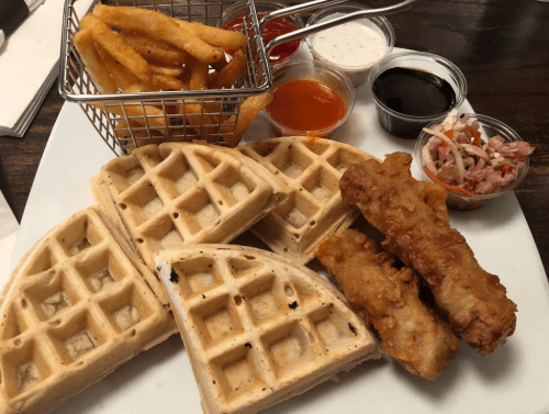 A plate with waffles, fried chicken, fries in a basket, and various dipping sauces.