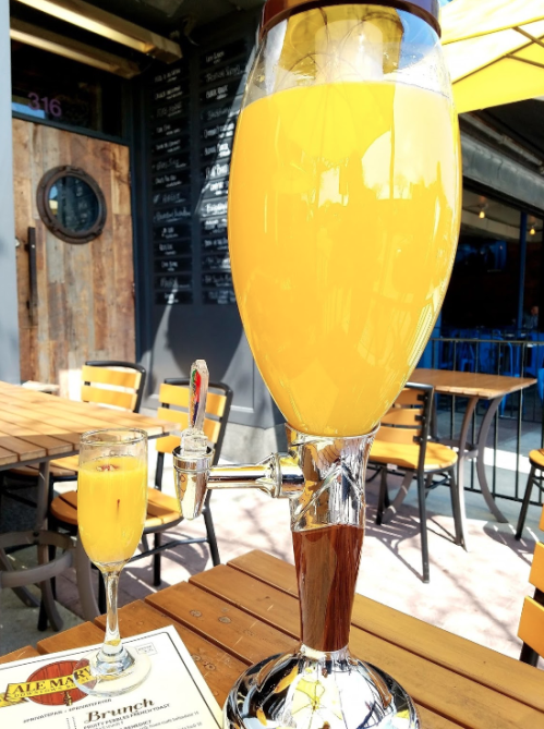 A large glass of orange drink with a tap beside a smaller glass, set on a wooden table outdoors.