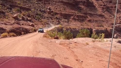 A dusty dirt road winds through rocky terrain, with a vehicle driving in the distance amidst desert vegetation.
