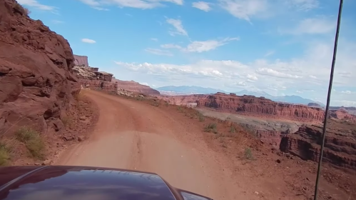 A dirt road winds through red rock formations under a blue sky with scattered clouds, showcasing a scenic landscape.