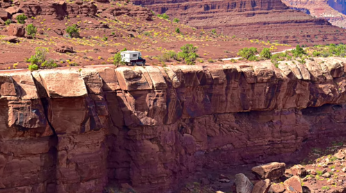 A camper parked on a rocky cliff edge, surrounded by red rock formations and sparse vegetation.