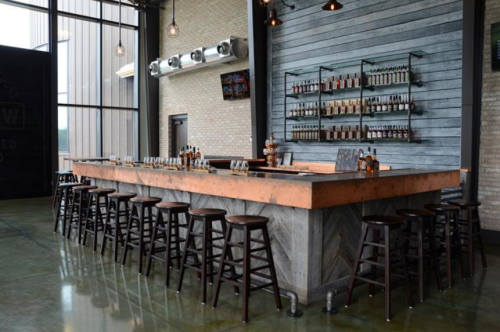 A modern bar with a copper countertop, wooden stools, and shelves lined with bottles against a brick wall.