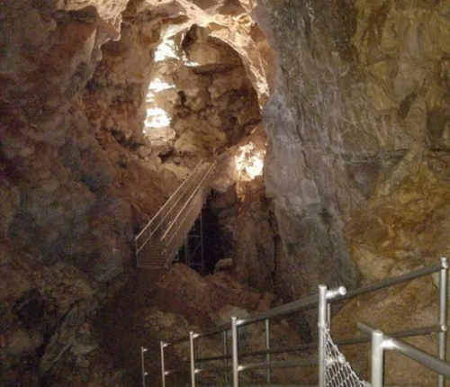 A dimly lit cave interior with rocky walls and a metal railing leading to a staircase.