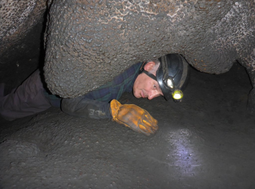 A person in a helmet and gloves crawls through a narrow cave passage, illuminated by a headlamp.
