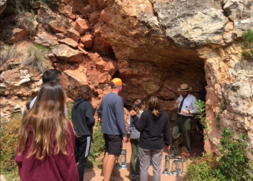 A group of people gathers around a park ranger at a cave entrance, surrounded by rocky terrain and greenery.