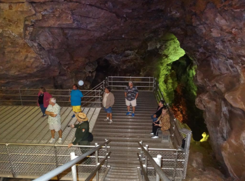Visitors explore a cave interior, standing on a metal platform surrounded by colorful rock formations.