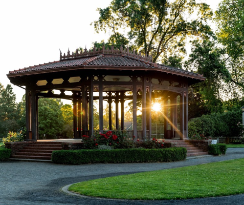 A wooden gazebo surrounded by greenery and flowers, with sunlight streaming through at sunset.
