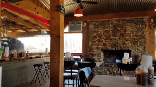 Cozy restaurant interior with wooden beams, stone fireplace, and tables, featuring condiments on the counter.