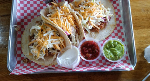 Two tacos filled with shredded meat, cheese, and toppings, served with salsa, guacamole, and sour cream on a tray.