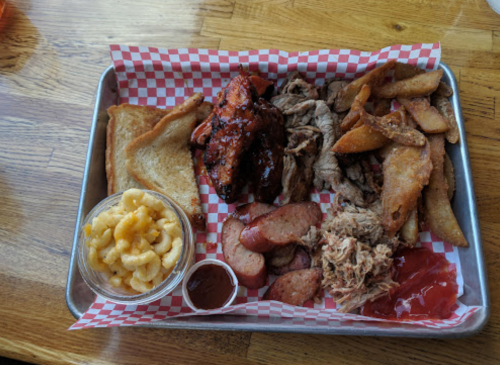 A tray of barbecue featuring chicken, pulled pork, sausage, fries, toast, and macaroni and cheese in a bowl.