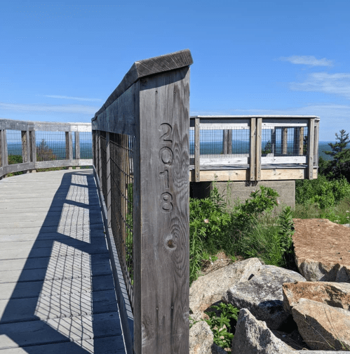 Wooden railing with "2018" carved into it, overlooking a scenic view of the ocean and greenery under a clear blue sky.