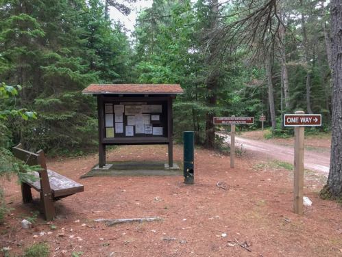 A wooden information kiosk in a forested area, with signs and a bench nearby, surrounded by pine trees and a dirt path.