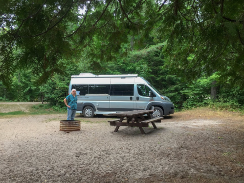 A person stands near a picnic table and fire pit, with a camper van parked in a wooded campsite.