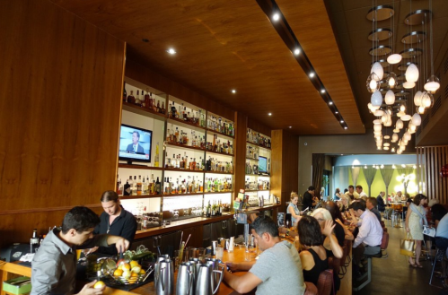 A modern bar with a wooden interior, shelves of liquor, and patrons enjoying drinks and food at the counter and tables.
