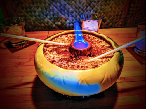 A colorful bowl filled with a flaming drink, garnished with chopsticks, set on a wooden table.