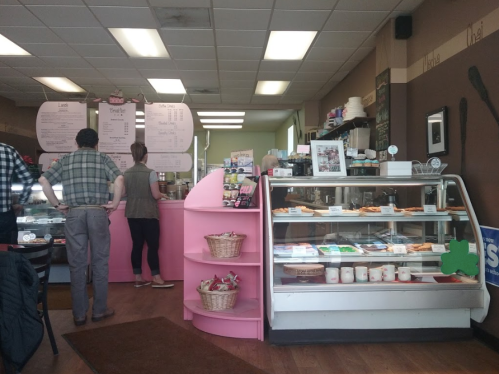 A cozy café interior with a pink display case, menu board, and customers ordering at the counter.