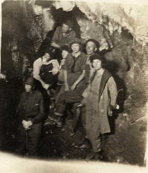 A group of six people poses inside a cave, surrounded by rock formations, in early 20th-century attire.