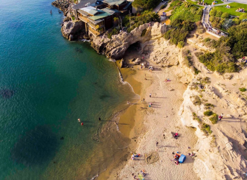 Aerial view of a sandy beach with people, clear water, and rocky cliffs, surrounded by greenery and buildings.